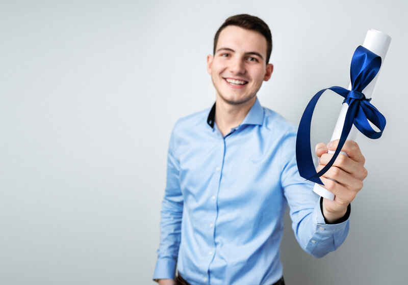 junger Mann hält zusammengerolltes Abschlusszeugnis mit blauer Schleife in der rechten Hand nach vorn